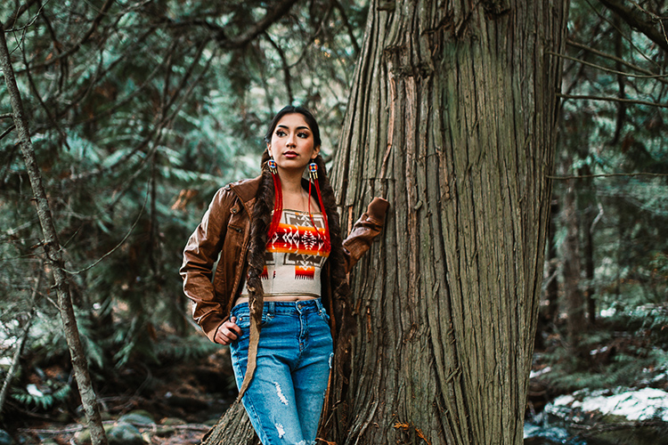 New student Sariel Sandoval, a Native American from a Montana reservation, poses for her high school portrait against a tree in the woods. She has long black braids and clothing with Native American designs.