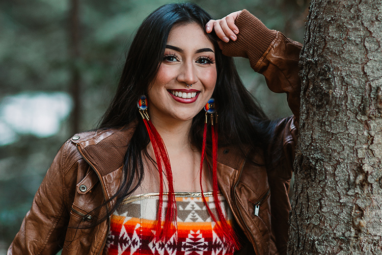 Sariel Sandoval poses for her high school portrait with long, flowing black hair, clothing with Native American designs, and leans against a tree in the woods.