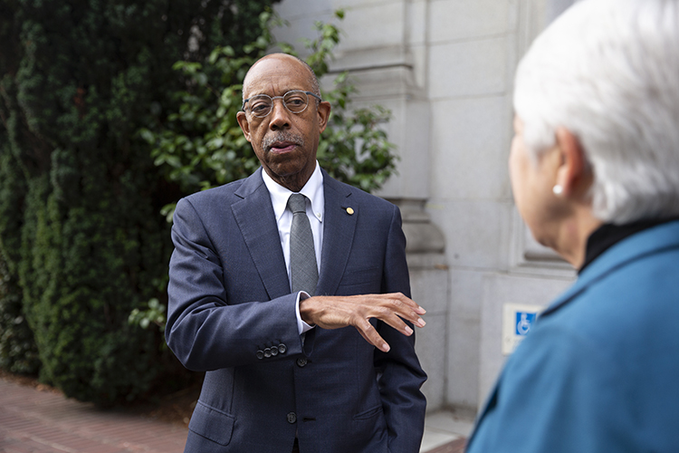 drake, in a suit, gestures whiile talking to chancellor carol christ outside of california hall