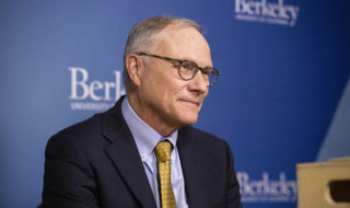 card, in a suit, sits in front of a berkeley branded background