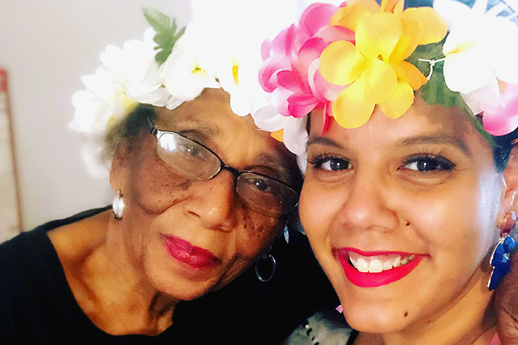 matos, wearing a flower around her head, poses with her grandma, who has glasses and is also wearing a flower hat