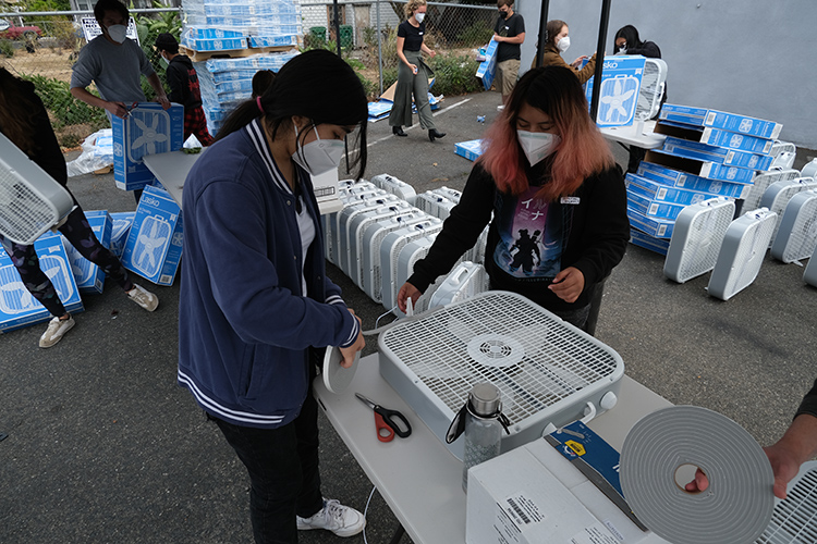 Two volunteers work under a tent at a Common Humanity Collective event and place foam weather stripping along the perimeter of a box fan's frame. This creates a tight seal between the fan and filter to reduce leaks that would allow dirty air to bypass the filter.
