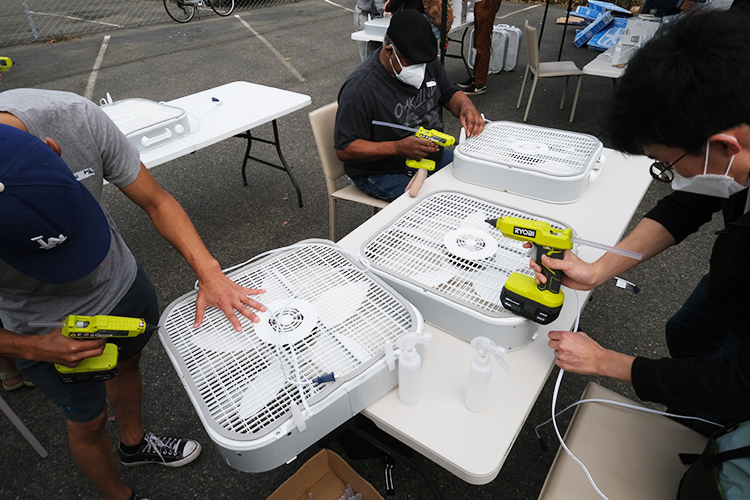 Three volunteers at a table use hot glue guns to secure the foam weatherstripping to a box fan, making a DIY air purifier. This allows the stripping to be able to withstand future filter changes.