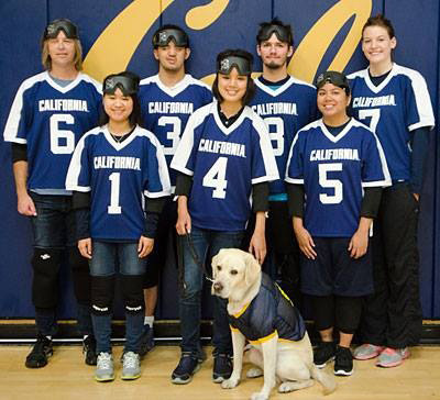 Ann Wai-Yee Kwong smiles for a group photo with members of Berkeley's goalball team. Each team member wear a blue and white Cal jersey with a number on it. A dog wearing a jersey poses with the team as well.