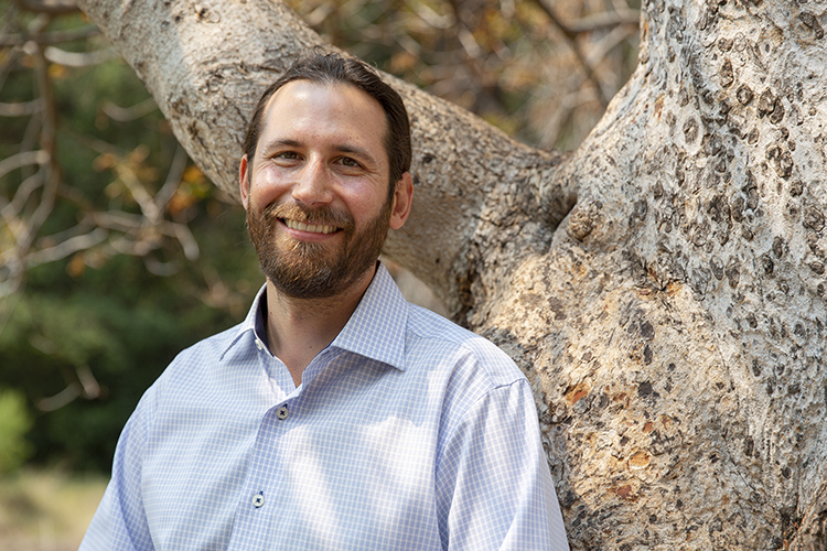 New faculty member Peter Nelson leans against a tree in Faculty Glade and smiles. He has a short pony tail and a beard and is a Native Californian.
