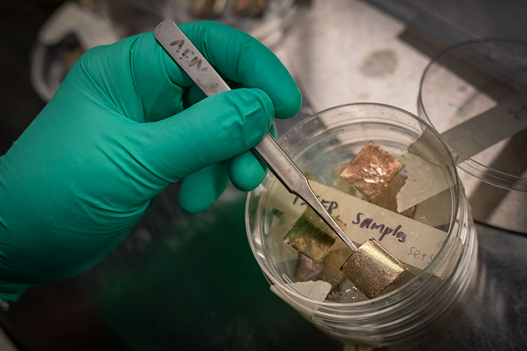 A photo shows a gloved hand holding a small metal sheet using a pair of tweezers