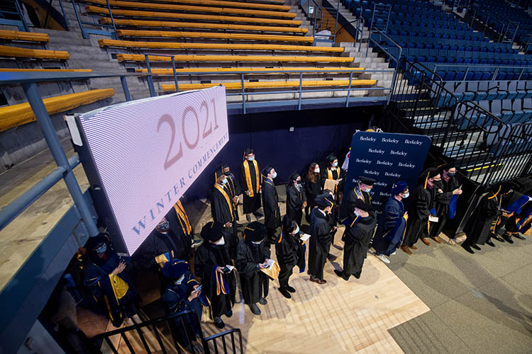 a crowd in graduation gear stands under a sign that says 2021 commencement