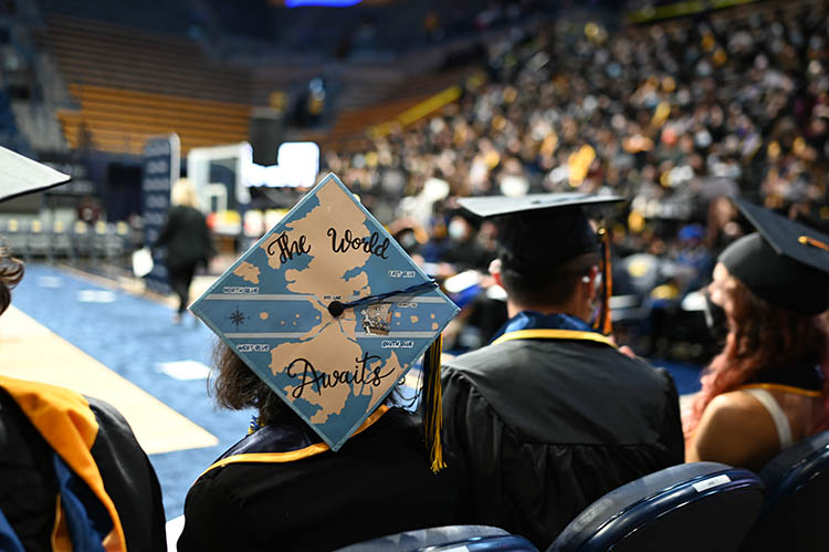 a graduate wears a hat that says the world awaits