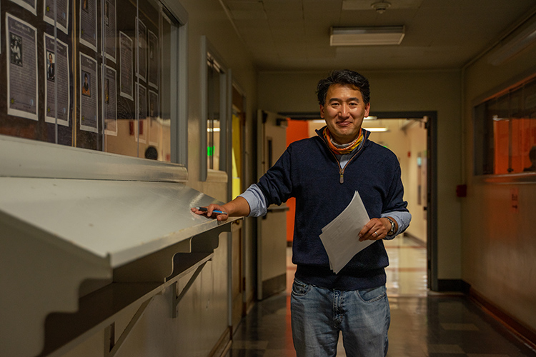 Professor Eugene Chiang, who teaches astronomy and earth and planetary science, stands in a hallway at Berkeley, smiling at the camera and holding some paperwork.
