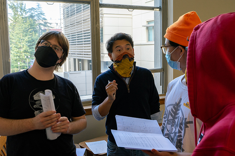 Professor Eugene Chiang, wearing a gold bandana over his mouth during the COVID-19 pandemic, talks with masked students in a small group, all of them standing up, in his Order of Magnitude Physics course.