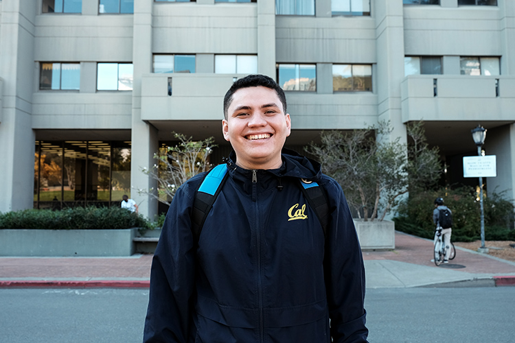 Ivan Chavez, a Berkeley third-year student major in history, stands outside Evans Hall smiling and wearing a Cal jacket and a backpack.