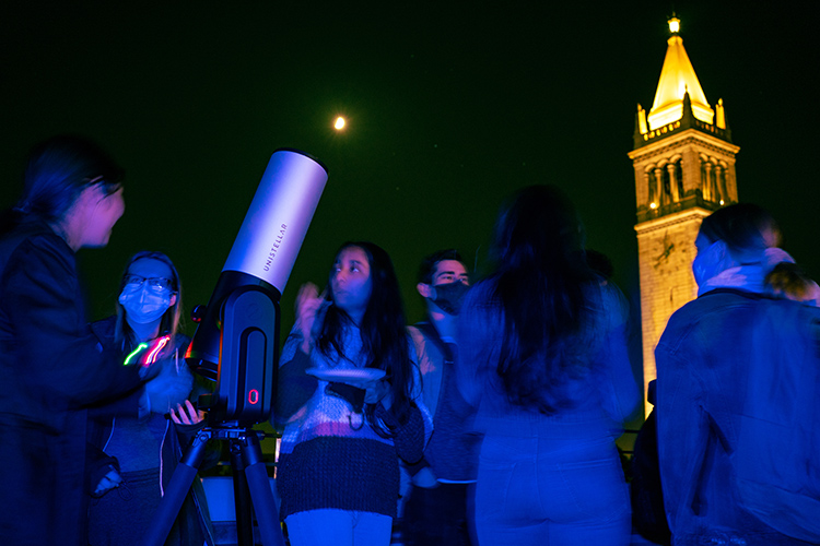 Students with telescopes gather at night on Memorial Glade to look at the heavens at a star party. The Campanile is lit up in the background.
