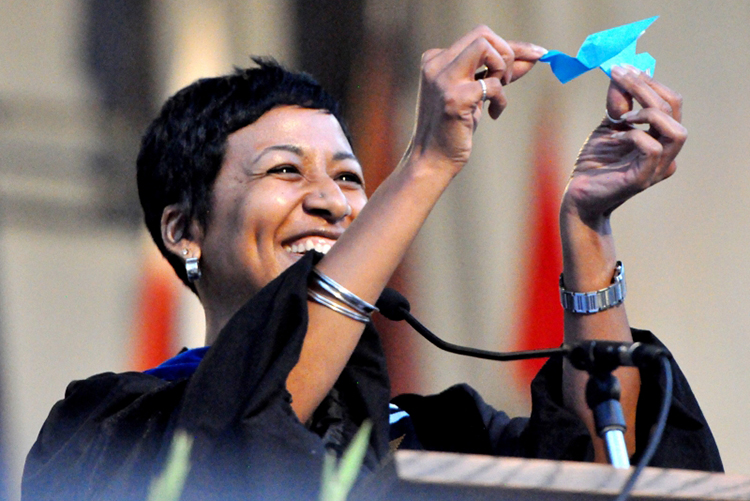 Amani Allen smiles at a graduation podium while holding up a blue origami crane.