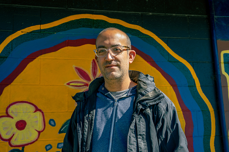 Ari Neulight, a campus social worker who does outreach to the homeless on and near campus, poses next to a mural at People's Park near campus. He has a serious look on his face and is wearing eyeglasses, a jacket and a zip-up sweatshirt.