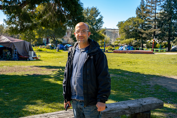 Ari Neulight, the campus's homeless outreach coordinator, poses for a photo in People's Park. He's wearing eyeglasses, jeans, a sweatshirt and jacket and holding a cellphone.