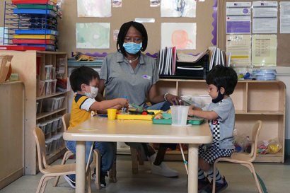 a child care teacher sits between two young children at a tiny indoor table as the children focus on learning materials