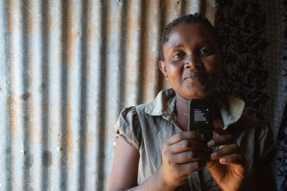 A photo of a woman standing against a wall made of corrugated metal, holding up a cell phone. The screen of the phone reads, "You have received your GiveDirectly payment in the amount of 2280"