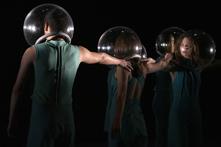 three dancers stand on a dark stage wearing bubbles over their heads with their hands on each others shoulders
