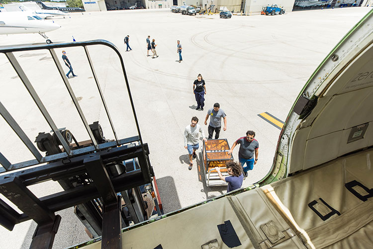 A photo taken from the cargo door of an aircraft shows a group of people pushing an orange box across the tarmac on a cart.
