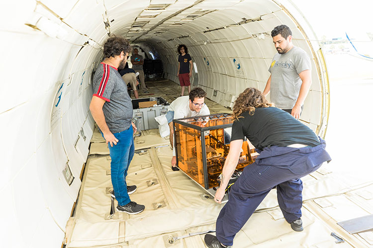 Two people lower a clear orange box to the floor of an aircraft, while a third person looks on. The inside of the aircraft has no seats and the floor is made of beige matting.