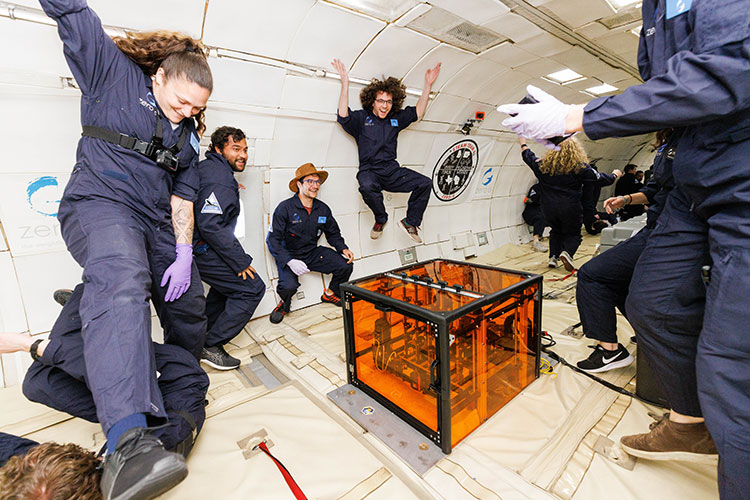 A group of people dressed in blue jumpsuits surround a clear orange box that is attached to the floor in the cabin of an aircraft. Many of the individuals are smiling and floating in the air because they are experiencing zero gravity.