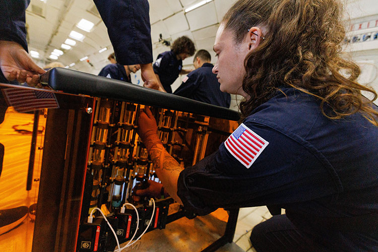 A person wearing a blue jumpsuit adjusts a component inside inside a clear orange box