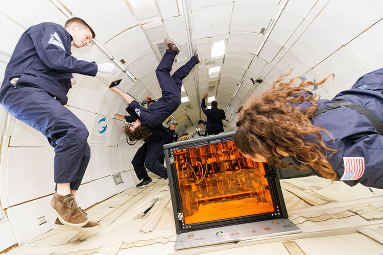 A person wearing a blue jumpsuit appears to be walking on the ceiling inside the cabin of an aircraft. In the foreground, two people, both floating in the air, look at a clear orange box that is secured to the floor.