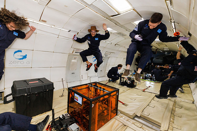 Three people wearing blue jumpsuits float near the ceiling within the cabin of an aircraft.
