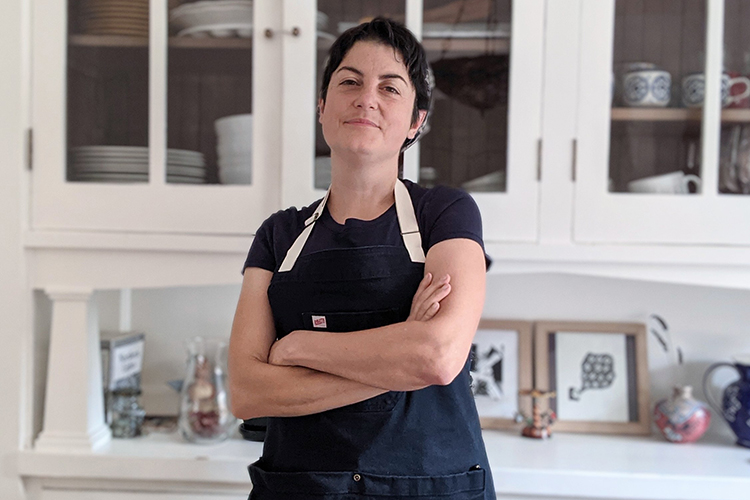 A photo of printmaker Irenka Pareto, standing with her arms crossed in front of her, an apron on and looking at the camera with a half-smile.