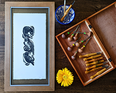 A woodblock print of falcons sits on a table along with artist Irenka Pareto's tools and some flowers.