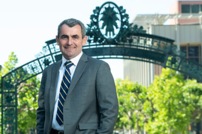 Oliver O'Reilly stands in front of Sather Gate wearing a suit and tie and smiling at the camera.