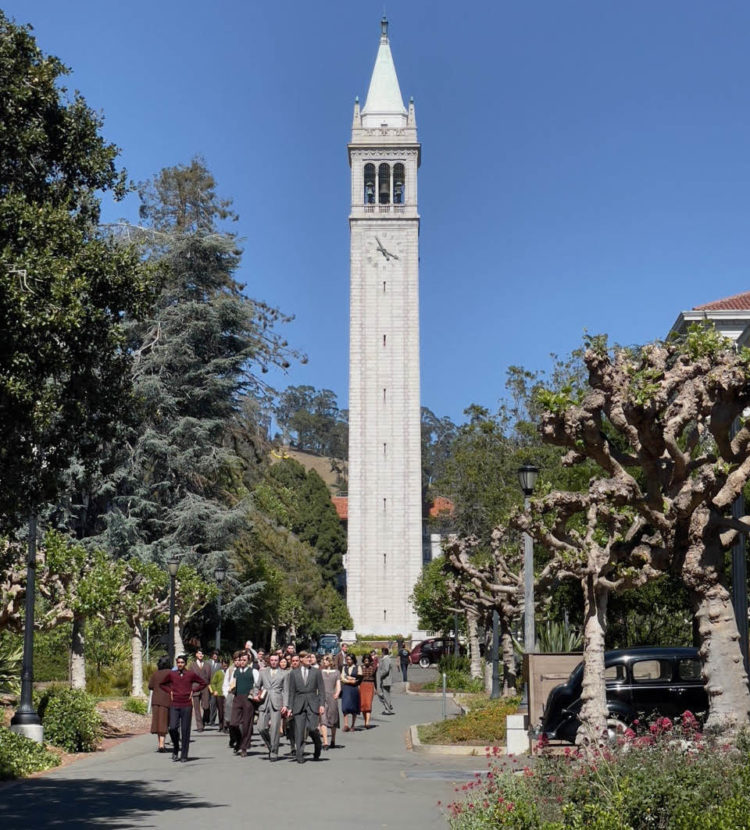 Cast members in period costumes walk near old cars and the Campanile.