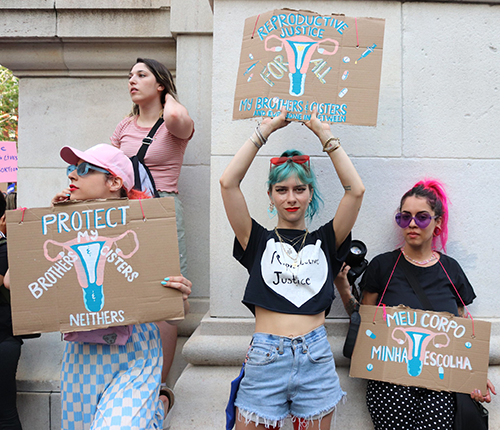 three people at a rally hold up signs for reproductive justice for all