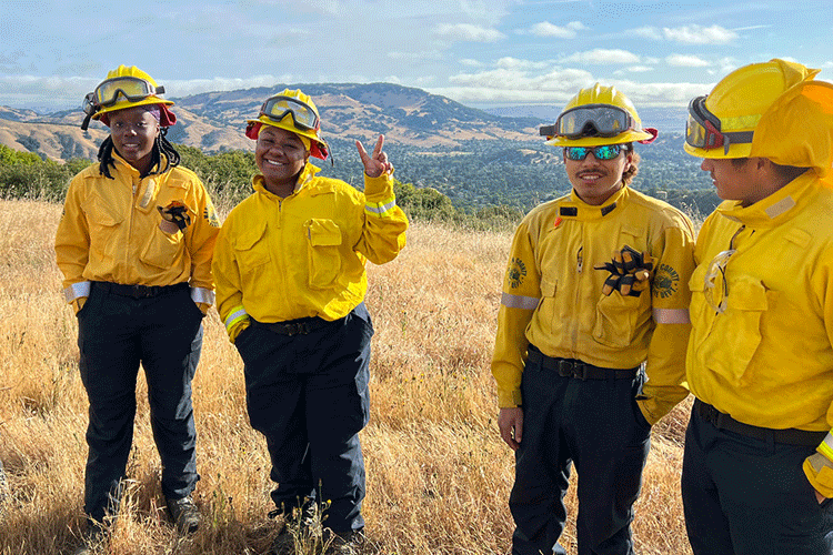 Four young adults wearing yellow shirts and hardhats stand in a grassy California field, with rolling hills in the background. One of the young adults holds up a peace sign to the camera.