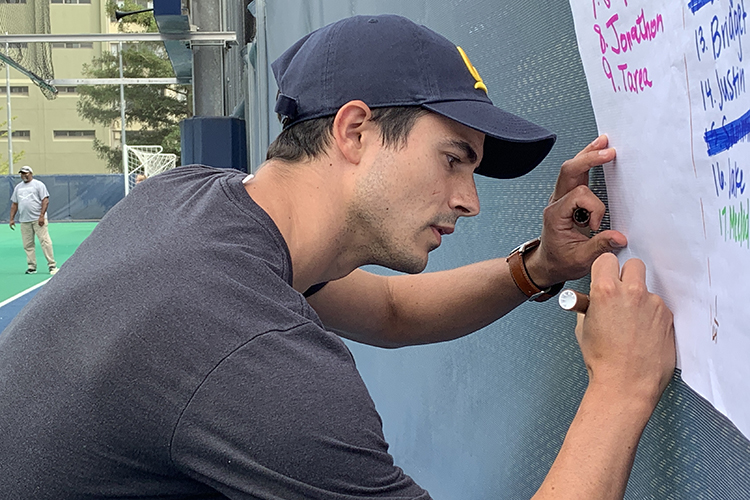 Wearing a baseball cap and a navy blue T-shirt, Jeffrey Brooks puts data on a large white piece of paper during Game 3 of the Student Affairs Summer Softball League's 2022 series.