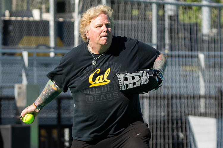 Stacy Hunter pitches the ball at the opening game of 2022 for the Student Affairs Softball League. She is an older woman with a Cal T-shirt and tattoos on her lower arms.