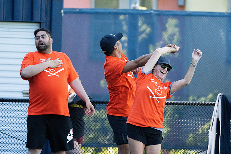 Three members of the Student Affairs softball league who play on the Dean of Student Centers team enjoy a laugh and celebrate during Game 1 of the season. Their team wears orange T-shirts.