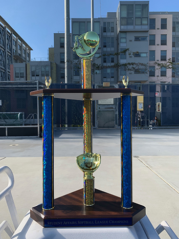 Here's the trophy for the team that wins the 2022 Student Affairs Summer Softball League championship game on Tuesday, Aug. 9, at 4:45 p.m. It's tall with a middle gold column and two side columns in blue. There are at least two metal mitts on the trophy.