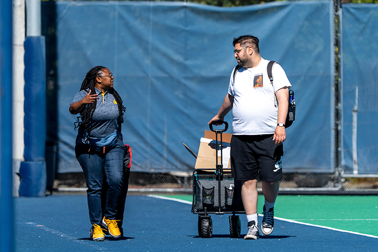 Two staff members walk down the edge of an athletic field, one of them is pulling a cart with supplies for the softball game they will be part of after work.