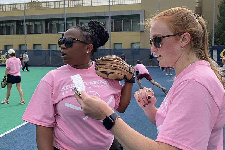 Wearing pink T-shirts, sunglasses and discussing their team's progress on the sidelines, co-captains Edtivia Rutherford and Bridget Bucey stand close together on the sidelines. Edtivia holds a mitt, Bridget holds a notepad.