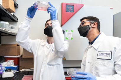 man and woman in face masks and white coats looking upward at Petri dish