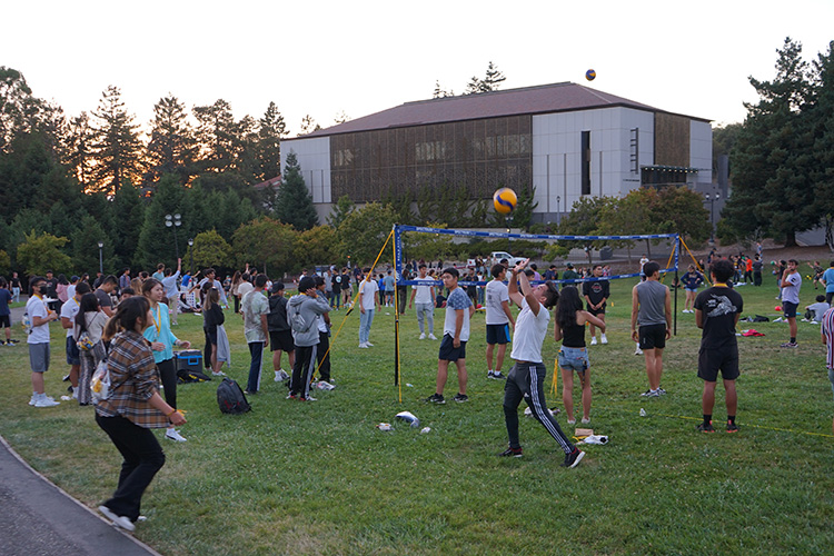 On Memorial Glade, dozens of incoming students relax and play volleyball as part of Golden Bear Orientation. Doe Library is in the background.