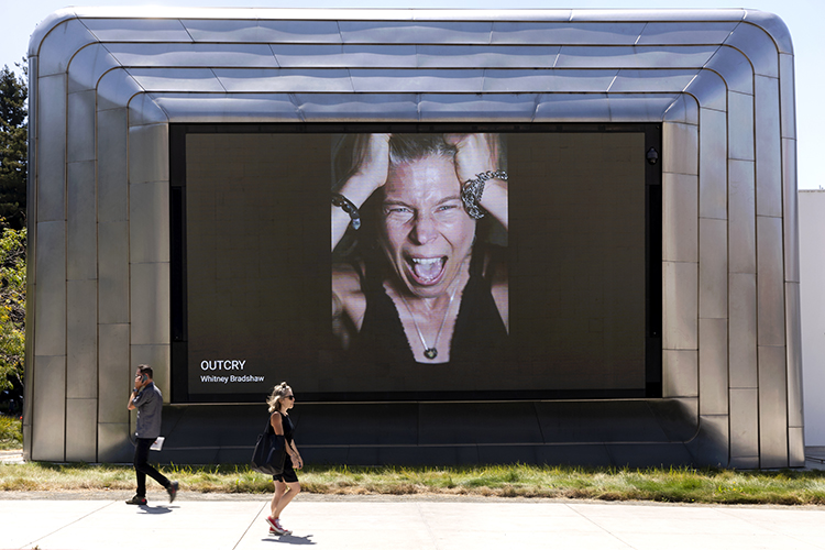 Two people walk past the giant screen on one side of the Berkeley Art Museum and Pacific Film Archive. The screen is displaying a photo of a woman screaming that is part of a collection of photos called OUTCRY by artist Whitney Bradshaw.