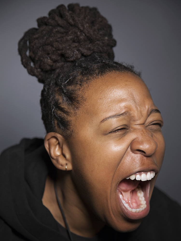 An African American woman screams at full volume in this photo from a collection called OUTCRY by photographer and social worker Whitney Bradshaw. The woman is young, is wearing a bun and has her eyes squeezed tight while she yells with her mouth open.