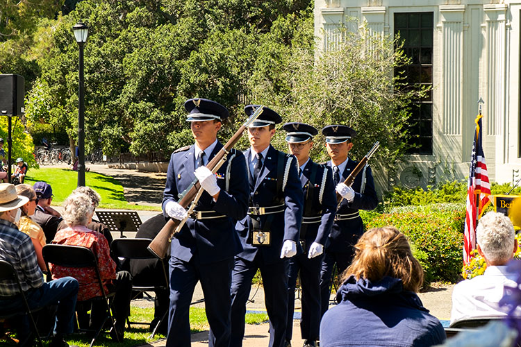 four cadets in blue uniforms march down the aisle carrying simulated weapons