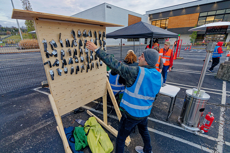 A researcher wearing a blue safety vest reaches for a grid of nearly 100 car keys that are hanging from a board. The researcher and the board are both outside in a parking lot.