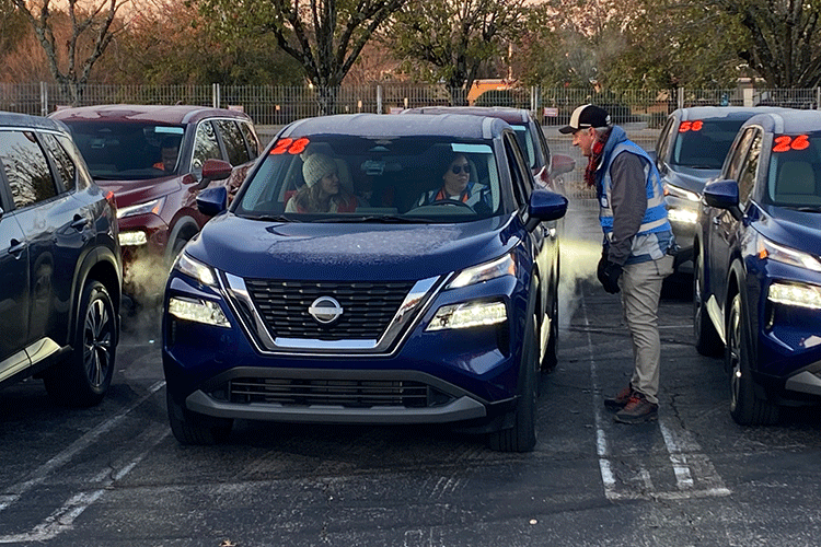 A researcher wearing a blue safety vest stands next to a blue Nissan Rogue vehicle, talking to a person who is sitting in the driver’s seat. A second person is sitting in the passenger seat.