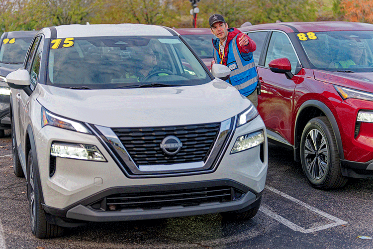 A researcher wearing a blue safety vest stands next to a grey Nissan Rogue vehicle, talking to a person who is sitting in the driver’s seat. The researcher is pointing to a location behind the camera.