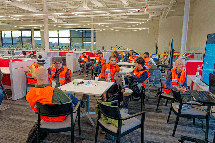 A group of people wearing a orange safety vests sit around tables in a large open office space.