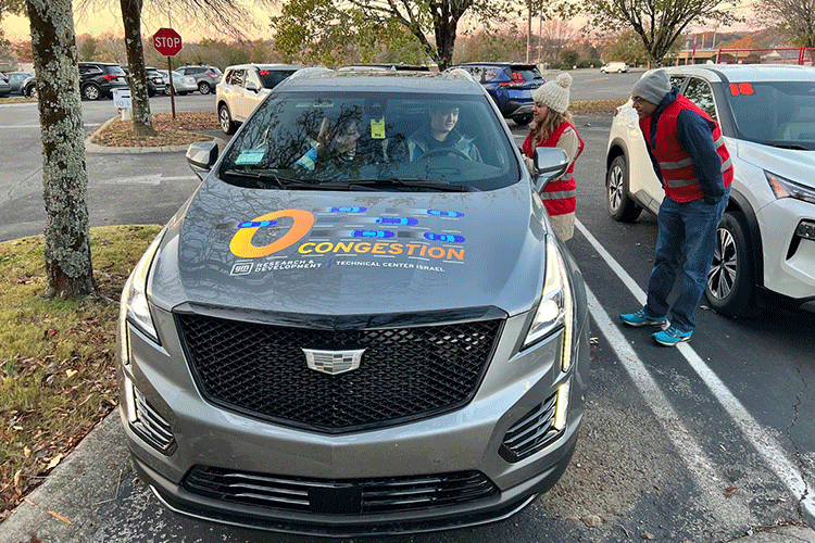 Two individuals wearing red safety vests stand next to a grey Cadillac XT5 vehicle, talking to a person who is sitting in the driver’s seat. A second person is sitting in the passenger seat.